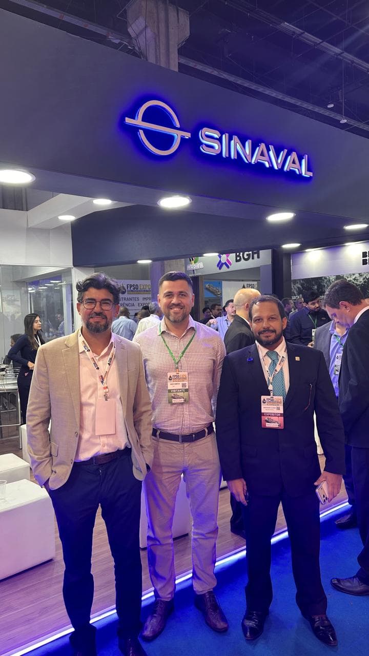 Three men in business attire posing in front of a SINAVAL booth at an expo.