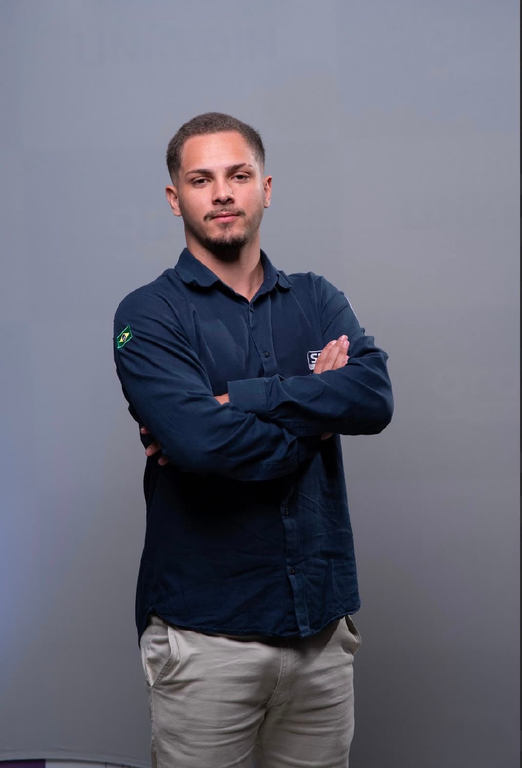 Man with crossed arms wearing a blue shirt with a Brazilian flag patch against grey.